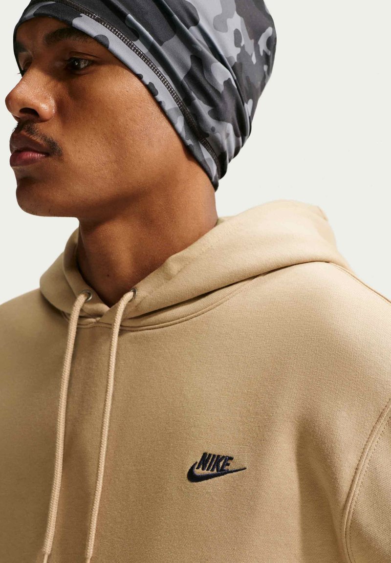 Mann trägt einen beigen Nike-Hoodie und eine graue Camouflage-Kopfbedeckung, seitlich blickend vor einem einfarbigen hellen Hintergrund.