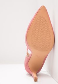Chaussure à talon haut en daim rose à bout pointu, inclinée vers le haut, montrant une semelle lisse couleur caramel avec un motif ondulé antidérapant et un talon fin.