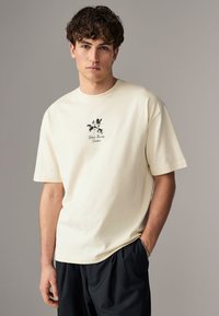 Off-white cotton t-shirt with short sleeves, featuring a small black floral graphic and text design at the chest. Relaxed fit, round neckline.