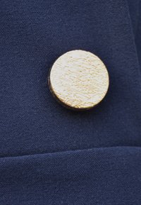 Złoty okrągły guzik o fakturowanej powierzchni, umieszczony na tle ciemnoniebieskiego materiału. Posiada gładką krawędź i subtelny połysk.