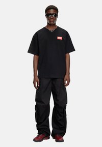 Sort oversized t-shirt med rødt "DIESEL" logo, parret med løse sorte cargo bukser og sneakers med røde accenter. Modellen har også solbriller på.
