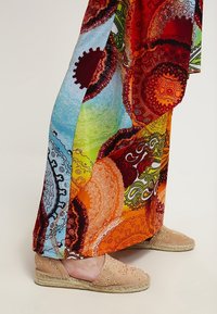 Jambes portant un pantalon large coloré avec des motifs circulaires et des espadrilles beiges avec de petites décorations sur un fond clair.