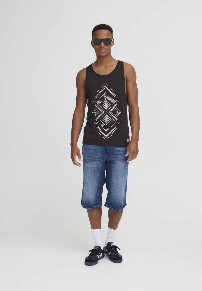 Black tank top with white geometric pattern, blue denim shorts, white socks, and black sneakers, accessorized with sunglasses.