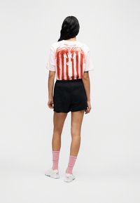 Λευκό μπλουζάκι με κόκκινες κάθετες ρίγες, με το κείμενο "NIKA" και τον αριθμό "1" στην πλάτη. Συνδυάζεται με μαύρο σορτς και ροζ κάλτσες.