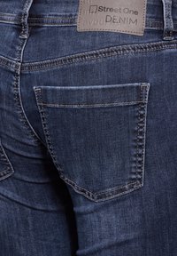 Mörkblå denimjeans med en bakficka. Har synliga sömmar och en läderetikett vid midjan med varumärkesnamn präglat.