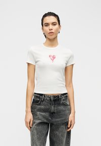 Λευκό, κοντομάνικο τοπ με γραφικό ροζ καρδιάς στο στήθος· διαθέτει κοντά μανίκια και ρεβέρ στο λαιμό. Φορεμένο με μαύρο, φθαρμένο τζιν.