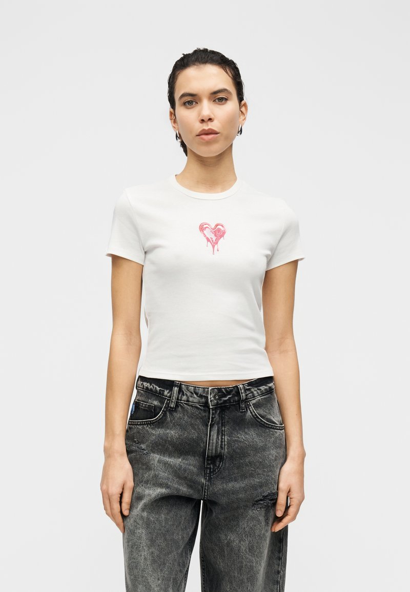 Λευκό, κοντομάνικο τοπ με γραφικό ροζ καρδιάς στο στήθος· διαθέτει κοντά μανίκια και ρεβέρ στο λαιμό. Φορεμένο με μαύρο, φθαρμένο τζιν.