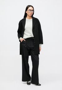 Melns pārizmēra mētelis, pelēks džemperis, melnas bikses ar šaurām svītrām un melni apavi. Nēsāts ar brillēm, taisni mati, neitrāla fonā.