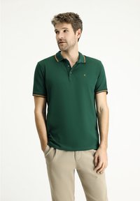 Polo shirt verde scuro con un accento arancione sul colletto e sulle maniche, realizzato in un tessuto liscio, dotato di una patta con due bottoni.