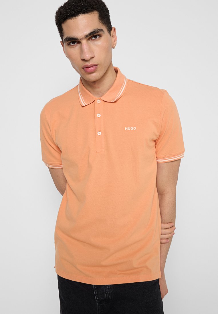 Koszulka polo z krókim rękawem w jasnopomarańczowym kolorze, wykonana z bawełny, z klasycznym kołnierzykiem z białym wykończeniem i małym białym logo "HUGO" na piersi.