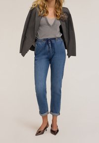 Jean en denim bleu à coupe décontractée, ourlets retroussés, associé à un haut argenté col en V et un blazer gris foncé à fines rayures. Chaussures noires à bouts pointus.