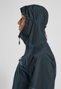Vandtæt hættejakke i mørk navy, med en lynlåslomme, justerbar hætte og tekstureret stof for holdbarhed. Sideprofilvisning.