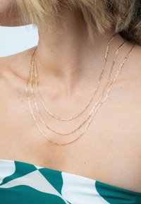 Goldbeschichtete Halskette mit drei Strängen, die strukturierte Glieder aufweist. An barem Haut getragen, harmoniert sie mit einem mehrfarbigen gemusterten Outfit.