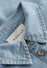 Γαλάζιο πουκάμισο από denim με άνοιγμα για το λαιμό, μονές ραφές σε καφέ χρώμα, δύο κουμπιά και λευκή ετικέτα ρούχου με την επιγραφή "NOELLA" στο εσωτερικό.