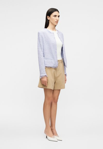 Γυναίκα με υφασμάτινο ανοιχτό μωβ σακάκι, λευκή εφαρμοστή μπλούζα, μπεζ σορτς μέχρι το γόνατο και λευκά μυτερά παπούτσια με χαμηλό τακούνι, που στέκεται πλαγίως.