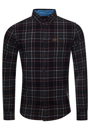 Homme debout contre un mur en bois portant une veste en cuir marron, une chemise à carreaux, un jean bleu et des baskets blanches avec des touches noires.