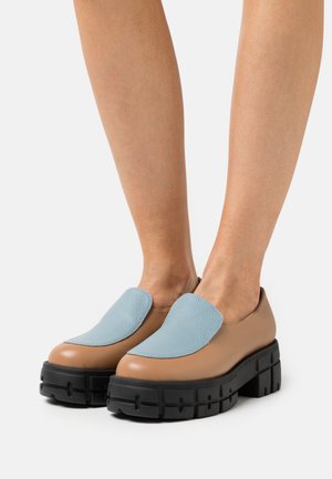 Zapatos marrones de piel tipo slip-on con un acento texturizado en azul claro, suelas de plataforma negras gruesas y diseño de punta redondeada.