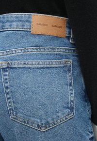 Jeans de ganga azul claro com uma etiqueta de marca em pele rotulada "SAM SøE". Apresenta um design clássico de cinco bolsos e detalhes de costura visíveis.
