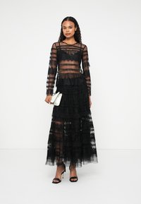 Rochie neagră din dantelă cu mâneci lungi, țesătură transparentă în straturi, textură ondulată și fustă fluidă; modelul ține o mică poșetă albă.