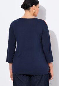 Top blu navy, ampio e con maniche a tre quarti, dettaglio con nodo laterale e accenti arancioni sulle spalle. Tessuto morbido, silhouette rilassata.