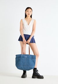 Denim toteveske med brune skinnhåndtak, med logo og synlig søm. Modellen har på seg en lys topp og et marineblått skjørt. Svart støvler.