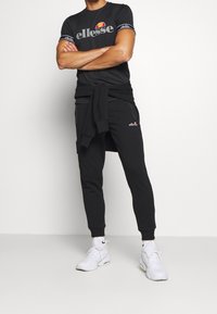 Černý sportovní outfit se slim tričkem s logem a kontrastními pruhovanými rukávy, doplněný o zúžené tepláky a bílé tenisky.