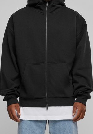 Man draagt een zwarte hoodie met ritssluiting en voorzakken over een wit shirt en lichtblauwe spijkerbroek tegen een grijze achtergrond.