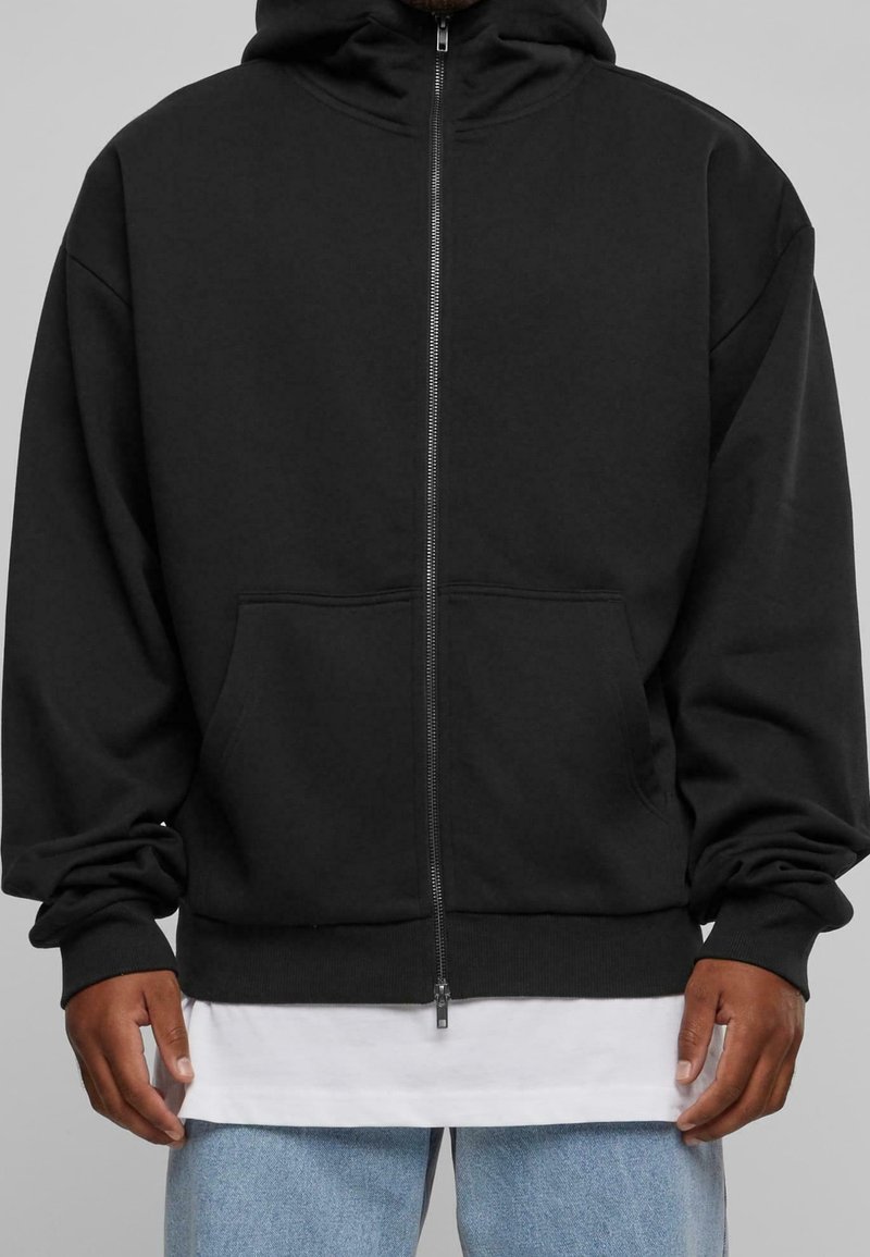 Homme portant un sweat à capuche noir zippé avec poches avant, par-dessus un t-shirt blanc et un jean bleu clair, sur un fond gris.