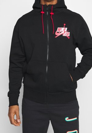 Sort lynlåstrøje med røde snore og rød Nike Air-logo på brystet, båret med sorte bukser med farverigt Nike swoosh-design.