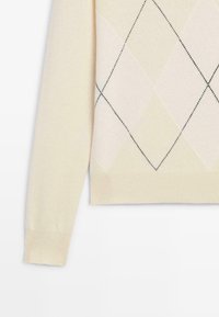 Pull beige clair avec un motif losange en deux tons de beige et noir. Il présente des poignets et un ourlet côtelés, une coupe raccourcie, et une texture douce.