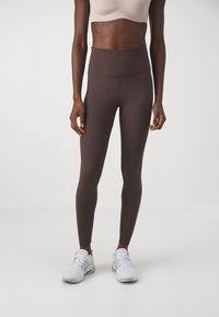 Hnědé legíny s vysokým pasem vyrobené z elastického materiálu s hladkou strukturou a bez viditelných vzorů, v kombinaci s bílými sportovními botami.