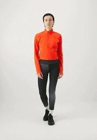 Inimene seisab seljas hele oranž lukuga pikade varrukatega topp, musta ja halli gradientiga legginsid, valged sokid ja mustad jalanõud ühetoonilisel taustal.