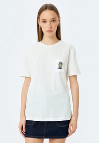 Witte katoenen t-shirt met korte mouwen, voorzien van een kleine geborduurde cartoonfiguur op de linkerkant van de borstzak, gecombineerd met een spijkerrok.