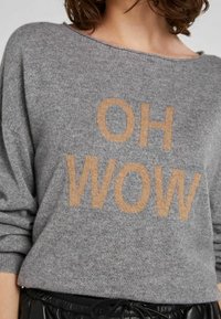 Donna che indossa un maglione grigio con le maniche arrotolate, con la scritta grande beige "OH WOW" e pantaloni neri con coulisse.