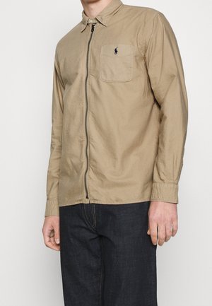 Chemise beige à manches longues avec fermeture éclair frontale et poche poitrine avec un petit logo sombre, portée avec un jean bleu foncé.