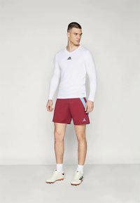 Camisola atlética de mangas longas brancas com logótipo preto, calções bordô com detalhes azul claro e sapatilhas de cor clara.