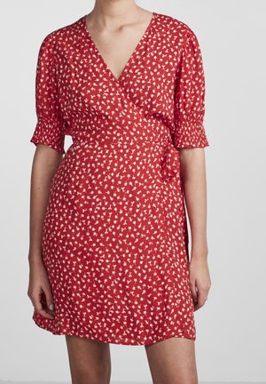 Femme portant une robe portefeuille rouge avec des motifs floraux blancs et des manches bouffantes courtes avec poignets froncés.