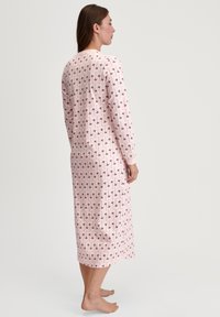 Vrouw die blootsvoets staat in een lang, mouwlengte nachthemd tot de enkel in het roze met een paars bloemenpatroon, zijwaarts naar links gekeerd.