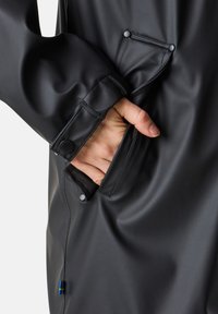Schwarze wasserdichte Jacke mit einer glatten Textur, ausgestattet mit einer Hand in einer Seitentasche und knopfbaren Manschetten für einen sicheren Verschluss.
