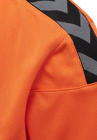 Orange atletisk trøje med tekstureret stof, kontrasteret af sorte skulderdetaljer med et zigzag-mønster i reflekterende materiale.