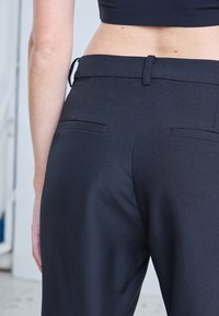 Gros plan d'une personne portant un pantalon noir taille haute sur mesure avec des poches arrière passepoilées et un haut court noir.