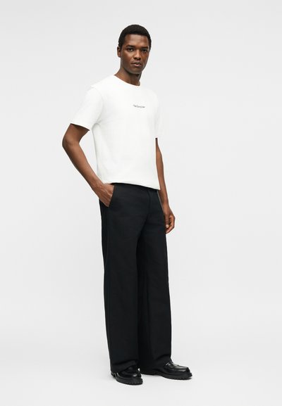 Άνδρας που φοράει λευκό κοντομάνικο μπλουζάκι και άνετο μαύρο παντελόνι με μαύρα παπούτσια, στέκεται με το ένα χέρι στην τσέπη.