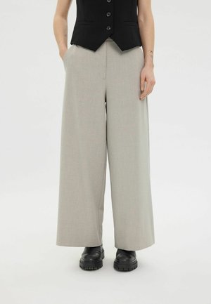 Personne portant un pantalon beige à jambes larges, un gilet noir sans manches boutonné et des chaussures noires épaisses, debout les mains dans les poches.