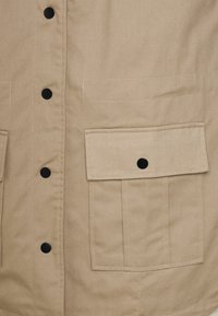 Casaco bege com botões pretos e dois bolsos frontais. Feito de um tecido durável; apresenta uma textura suave e um design simples e funcional.