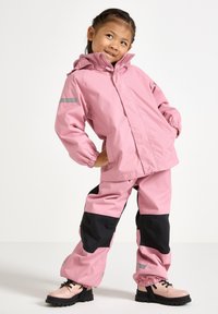 Rosa vattentät jacka med huva och elastiska ärmslut, kombinerad med matchande rosa byxor med svarta detaljer. Ljusa skor med svarta sulor.
