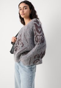Grauer Strickpullover aus einem strukturierten, flauschigen Material mit einem braunen Dekormuster. Kombiniert mit hellblauen Jeans.