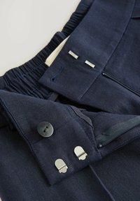 Tmavě modré kalhoty s texturovanou tkaninou, elastickým pasem, zapínáním na knoflík, dvěma kovovými klipy a postranní kapsou na zip.