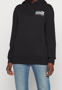 Sudadera negra con capucha que presenta un gráfico frontal con el texto "HUGO". Está hecha de tela suave con puños y dobladillo acanalados, y un diseño de ajuste relajado.