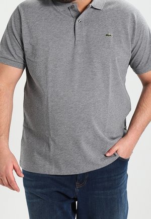 Homme portant un polo gris à manches courtes avec un logo crocodile et un jean bleu foncé, une main dans la poche et l'autre détendue.