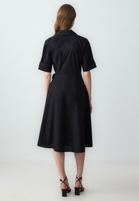 Robe noire à manches courtes, avec un col et une taille cintrée. Jupe évasée atteignant le milieu du mollet. Présente des détails de couture minimalistes et une texture lisse.